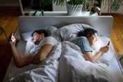 Paar mit T-Shirts bekleidet, liegt im Bett, sich gegenseitig abgewandt und schaut auf ihr Smartphone Paar mit T-Shirts bekleidet, liegt im Bett, sich gegenseitig abgewandt und schaut auf ihr Smartphone
